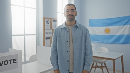 Fototapeta premium Handsome young hispanic man standing indoors with an argentinian flag in the background, emphasizing an electoral setting with voting booths and motivational materials around him