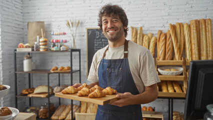 Young man in a bakery holding a tray of croissants with a smile surrounded by various baked goods