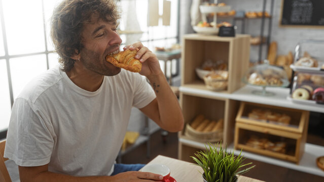 Young man enjoying a croissant in a cozy bakery with various fresh pastries displayed in the background - Powered by Adobe