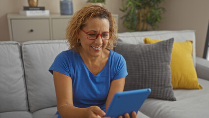 Mature hispanic woman with curly hair and red glasses, smiling while using a tablet in a cozy living room with a gray couch and yellow pillows.