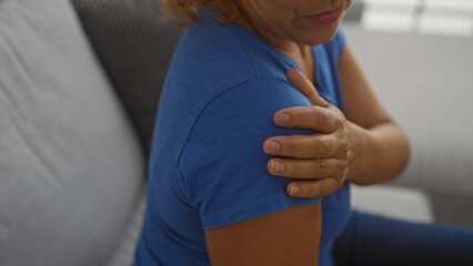 A woman holding her shoulder with her hand indoors, seated in a living room, wearing a blue shirt, suggesting pain or discomfort in her arm.