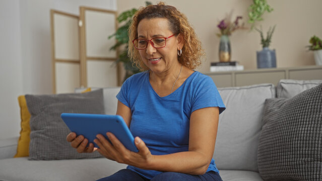 A mature hispanic woman with curly hair wearing glasses and a blue shirt uses a tablet while sitting comfortably on a sofa in her modern living room.