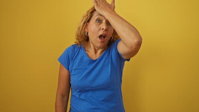 A middle-aged, hispanic woman with curly hair and a surprised expression touches her forehead in disbelief, isolated against a yellow background.