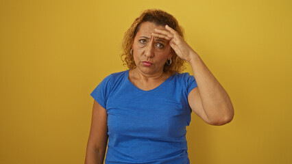 A middle-aged hispanic woman in a blue shirt with curly hair stands isolated against a yellow background, looking concerned and touching her forehead.