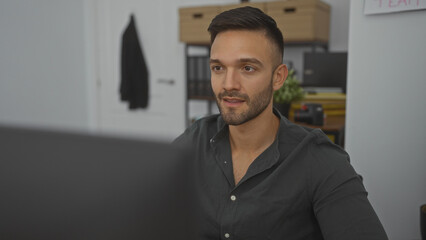 Obraz premium A young hispanic handsome man with a beard is sitting in an office, focusing intently while working indoors.