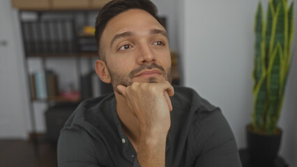 Handsome young hispanic man with a beard, sitting indoors in a thoughtful pose, in an office room...