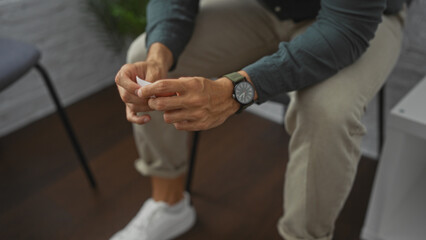 A hispanic man waits in an indoor room, seated and holding a paper, with his hands prominently displayed, evoking a sense of anticipation.