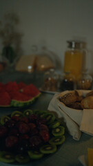 Assorted breakfast items including kiwi, plums, rolls, and orange juice arranged on an indoor table for a cozy morning meal