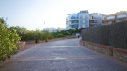 Blurred view of an outdoor pathway with greenery, defocused buildings and a distant person adding depth to a serene urban setting
