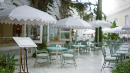 Blurred view of an elegant outdoor terrace with bokeh effect, featuring luxury dining chairs and tables under large umbrellas in a sophisticated restaurant setting.
