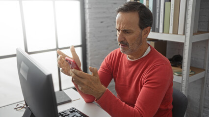 Fototapeta premium Man in office using smartphone looks puzzled seated at desk in modern workplace interior wearing red sweater near computer.