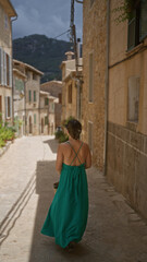 Young woman in green dress exploring the charming narrow streets of valldemossa, a picturesque village in mallorca, spain, surrounded by rustic stone buildings and mountains.