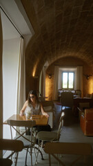 Young woman sitting alone at a wooden table in a dimly lit hotel cafeteria with rustic arched ceilings, wearing sunglasses and a light blouse, enjoying a moment of solitude by the window.