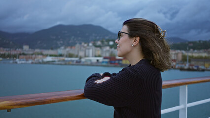 Woman enjoying scenic view from cruise ship dock with mountains and ocean in background, wearing sunglasses and black sweater, relaxed and smiling.