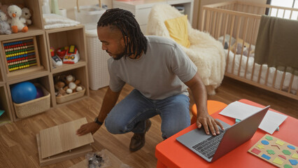 Young man with braids assembling furniture in a cozy bedroom with baby cradle nearby, surrounded by toys and using a laptop on a colorful desk.