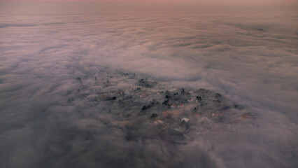 Aerial view captures a small town partially obscured by a thick blanket of mist during a serene sunrise, creating a mystical atmosphere