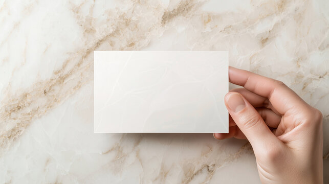 A hand with a slightly extended index finger delicately holding the corner of a blank transparent business card, set against a light marble background. The smooth texture of the ca