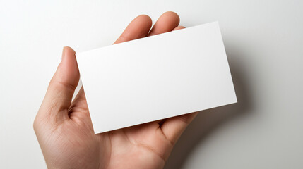 Obraz premium A blank white business card is held at a slight angle in the palm of a hand, set against a bright white background with soft ambient light. The smooth, glossy texture of the card c