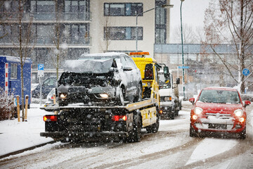 Tow truck carries damaged car during snowy city street scene.  Heavy snow falling.