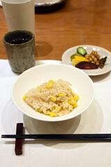 A bowl of steamed rice mixed with sweet corn is served alongside a small plate of colorful Japanese pickles, including cucumber, radish, and beans, creating a balanced and traditional meal.