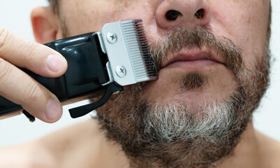 Close-up of a man trimming his thick, graying beard with an electric hair clipper, focusing on precision grooming and facial hair care.