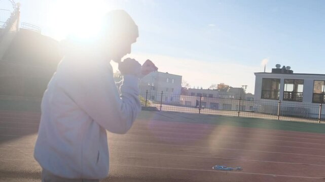 Man in hoodie boxing outdoors on running track in the bright sun. Workout at the stadium