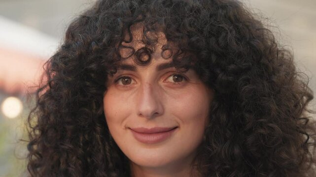 Very close portrait view of beautiful Caucasian woman's face. Girl looking at camera with focused expression. Gently smiling. Long curly hair. Cute brown eyes. Enjoying shadow from big umbrella.