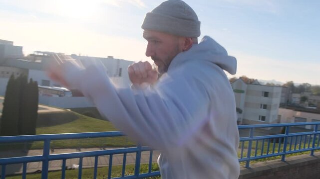 Man in light hoodie shadow boxing outdoors with blue railings and urban backdrop. Active and focused training