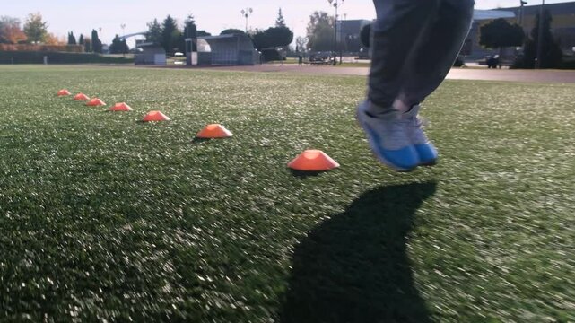 Close up of feet during football training. Athlete in grey pants and blue sneakers, navigating around orange cones on artificial turf