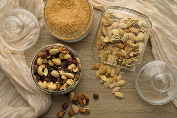 Top view cane brown sugar, nuts mix, raisins and scattered pumpkin seeds in transparent glass jars on wooden table, beige cloth on the background