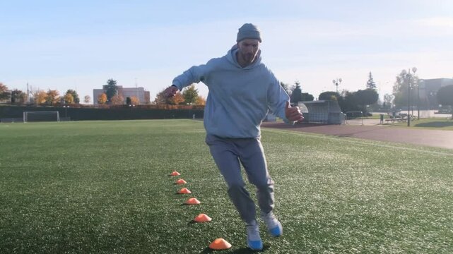 Footballer training. Man in blue hoodie, purple pants drills with orange cones on grass field. Sunny day, agility workout