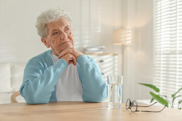 Fototapeta premium Thoughtful senior woman feeling lonely at table with glass of water indoors