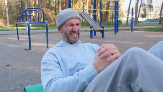 Man doing crunches, working hard on mat outdoors. Grey hat, blue sweatshirt, grey pants. Sport, exercise, fit, workout