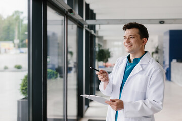 Young handsome doctor posing with a patient’s chart and pen by the window