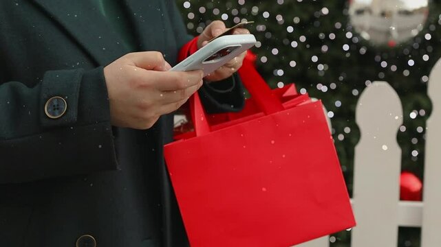 Woman in winter clothes holding credit card and smartphone while shopping for a winter holidays at shopping mall. Standing with gift box near just bought Christmas tree outdoors. 