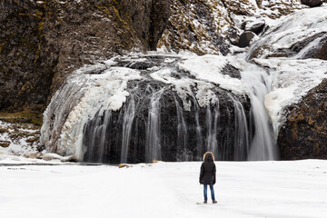 Traveler admiring frozen winter waterfall in ice-covered landscape. Adventure and cold-weather exploration highlighting nature&rsquo;s raw power.
