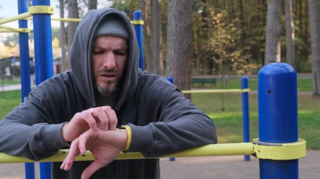 Athlete checks his watch at an outdoor gym. Man in hoodie is monitoring his workout progress and time. Fitness and sport