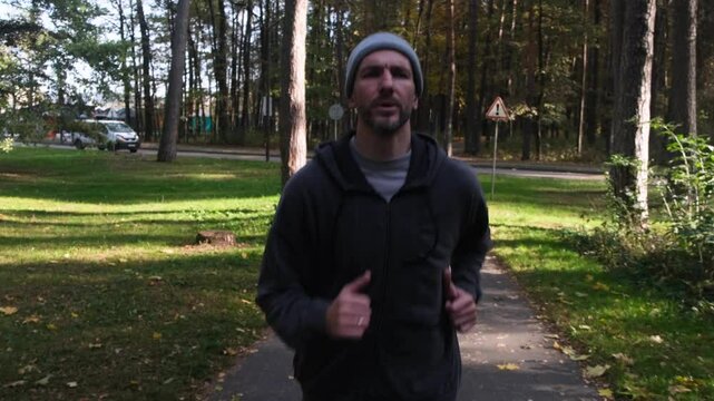 Man jogging in park. Close-up of face. He&rsquo;s in grey hoodie, pants and hat. Sunny day