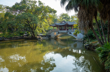 Naklejka premium A classic Chinese garden in a public parkland, features verdant trees, a graceful arched bridge, adorned with traditional architecture, spans a tranquil pond. Liuhou Park, Liuzhou, China.