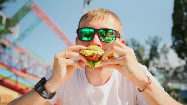 Portrait of a guy in sunglasses, happily eating a tasty burger with roller coasters in the background. The vibrant sky and thrilling rides create a festive summer atmosphere at the amusement park.