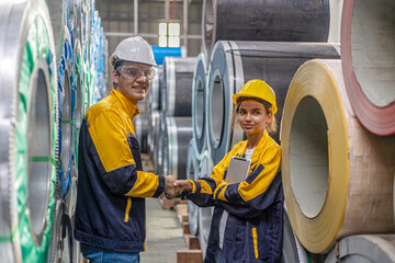 Two young engineers or mechanical workers in safety suit discuss and consult each other for their work routine next to the rolls of metal sheets in a factory. Partnership among two workers.