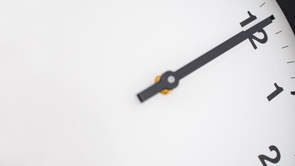 A close-up of an analog clock face focuses on the minute hand pointing at the 12, set against a minimalistic white background with black numbers.