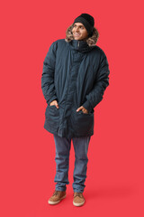 Young man in stylish puffer jacket and hat on red background
