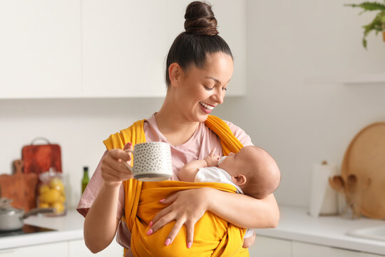 Young happy mother with little baby in sling and cup of coffee at kitchen