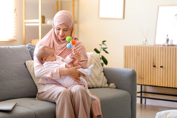 Happy Muslim mother in hijab and cute little baby playing with rattle at home