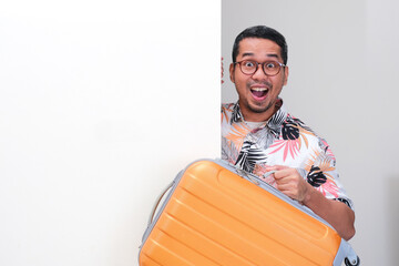 A tourist peeking behind a wall carrying suitcase with excited expression