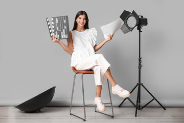 Young happy female film director with movie clapper, script and studio lighting sitting on chair...