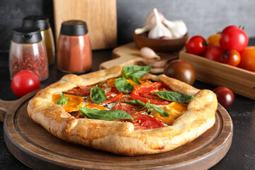 Wooden board of tasty tomato galette with basil on black background