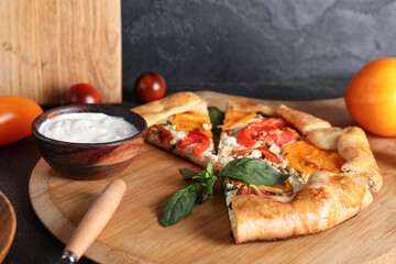 Wooden board with pieces of tasty tomato galette and bowl of sauce on black background
