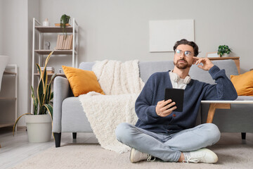 Thoughtful young man with e-reader sitting on floor at home
